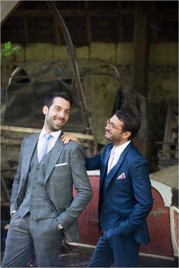 Portrait of two men, likely the groom and a groomsman or a same-sex couple, posing in front of a weathered red vintage piece of farm or industrial equipment inside a rustic barn or outbuilding. The man on the left wears a grey three-piece suit with a light blue patterned tie and white pocket square, while the man on the right wears a navy two-piece suit with a pink tie and pink pocket square and glasses. The man on the right has his hand resting on the other's shoulder, and both are smiling. The setting has a rustic, vintage aesthetic with aged wooden beams and distressed surfaces visible in the background. Medium portrait shot with a shallow depth of field.