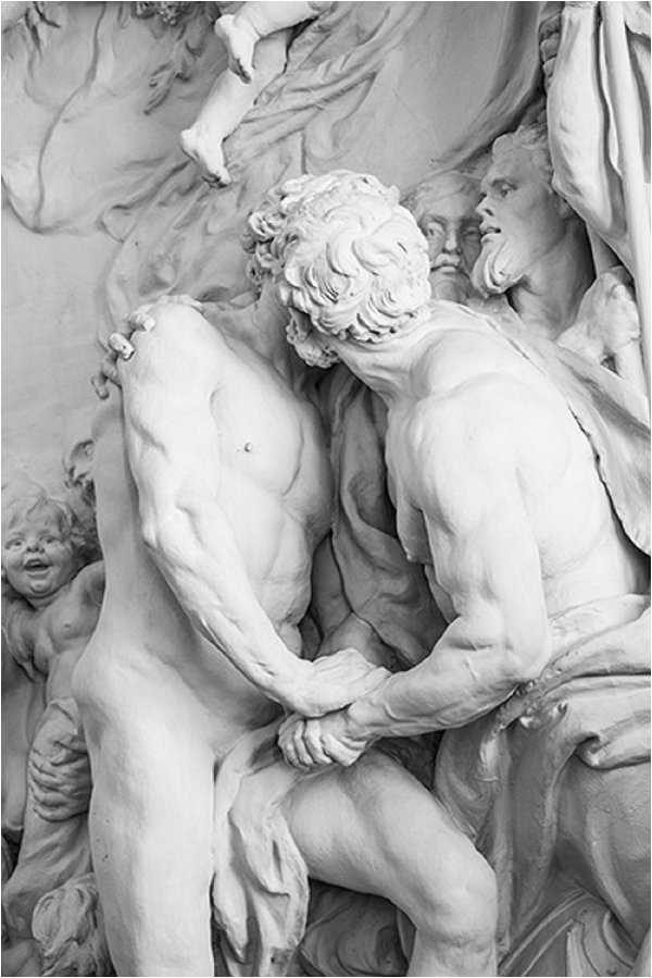 This image does not appear to be a wedding photograph. It shows a close-up of a classical white marble sculptural group featuring two muscular male figures in an embrace or struggle, with additional figures visible in the background including a bearded elder face and a cherub. The image is black and white, rendered in bright whites and soft mid-tones with minimal shadow contrast. This appears to be a detail shot of Baroque or Neoclassical architectural sculpture, likely from a French chateau or palace facade.