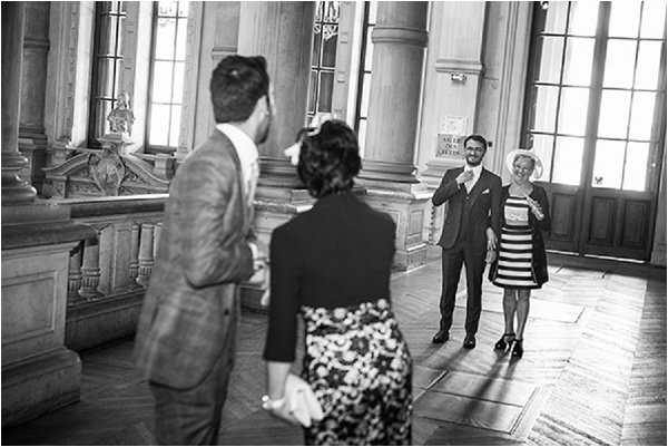 A black-and-white candid shot taken inside a grand civic or municipal building, likely a French mairie (town hall), featuring tall columns, large arched windows, and ornate parquet flooring. Four people are visible: a man in a plaid suit jacket stands with his back to the camera in the foreground, a woman in a floral dress faces away from camera beside him, while a groom in a dark suit and tie and an older woman in a short striped dress approach them from the background. The scene appears to capture a pre- or post-civil ceremony moment, with guests or family members greeting one another. The image is a wide candid shot with medium contrast, emphasizing the architectural grandeur of the interior space.