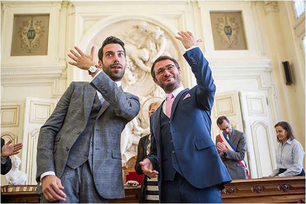 Two grooms celebrate their civil ceremony inside a French mairie (town hall), both raising their arms in a joyful gesture. The groom on the left wears a light grey textured tweed suit, while the groom on the right wears a navy three-piece suit with a pink tie and a pink pocket square. The interior setting features ornate classical architecture with white sculpted relief panels, decorative moldings, and the 'RF' (République Française) insignia visible on the walls. Several guests and an official are visible in the background, clapping. The shot is a mid-range portrait taken at a slight low angle, emphasizing the celebratory mood of the moment.