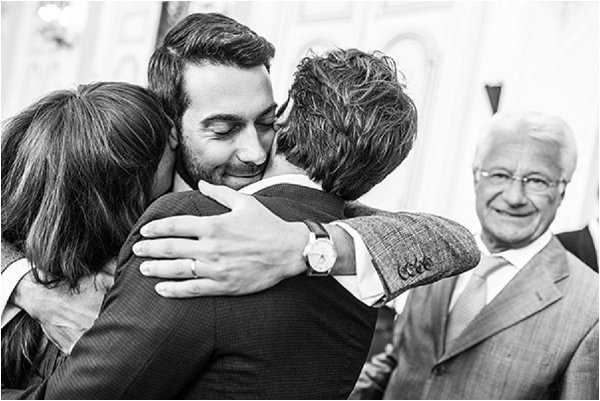 A black-and-white close-up portrait shot capturing a congratulatory moment, likely following a wedding ceremony, where a groom in a dark suit and watch embraces two guests simultaneously — a woman on his left and a man in a tweed jacket on his right. An older gentleman in a suit and glasses smiles warmly in the background. The image has strong contrast with bright highlights and deep shadows, giving it a candid, documentary feel.