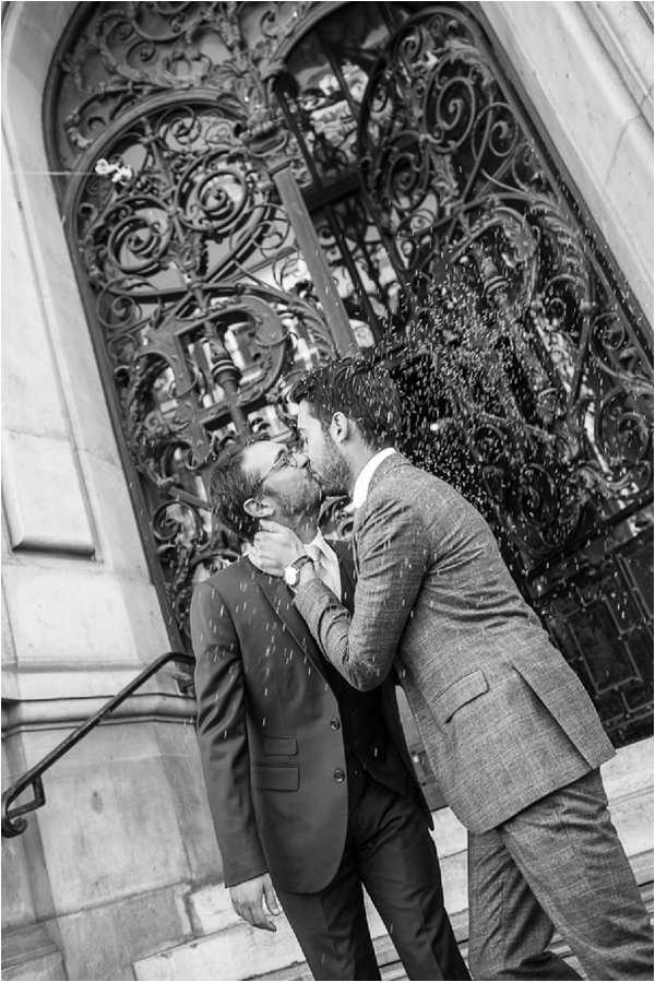 Black-and-white portrait of two grooms kissing on the steps outside an ornate wrought-iron arched doorway, likely at a mairie or historic civic building. Rice or confetti is being thrown over them, caught mid-air in the image. One groom wears a dark three-piece suit with a light tie and a watch visible on his wrist, while the other wears a textured light-grey suit. The composition is a mid-range portrait shot with strong contrast between the dark ironwork scrollwork of the door and the lighter stone architecture framing the couple.