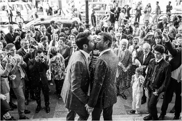 A black-and-white image capturing two grooms sharing a first kiss on the steps of what appears to be a French mairie (town hall), surrounded by a large crowd of approximately 40–50 guests who are throwing confetti and cheering. Both grooms wear dark suits with ties, and they are holding hands during the kiss. The confetti is visible mid-air, creating strong contrast against the dark suits in the high-contrast B&W tones. The composition is a wide shot taken from a slightly elevated angle, placing the couple at the center foreground with the crowd fanning out behind them and a street scene visible in the background.