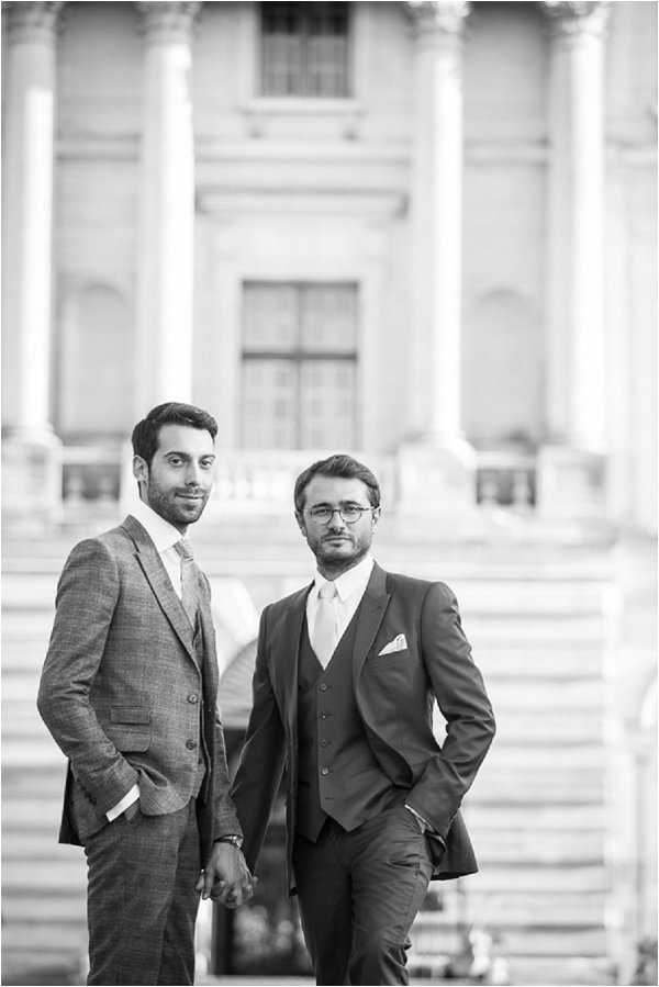 Black-and-white portrait of two grooms holding hands and standing together in front of a grand neoclassical building with large columns and wide stone steps visible in the soft-focus background. One groom wears a light-toned checked suit with a tie, while the other wears a dark three-piece suit with a light tie and a white pocket square, and glasses. Both have their free hands in their pockets and face the camera with composed expressions. The image is a mid-length portrait shot with shallow depth of field, giving the architectural background a blurred, high-contrast appearance against the sharper foreground figures.