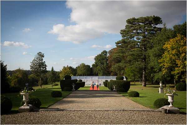 A wide-angle outdoor shot looking down a long gravel driveway lined with trimmed box hedges, ornamental stone urns, and manicured lawn toward a glass greenhouse or orangery structure in the background. Two figures, likely the couple, are walking along a red carpet runner laid over the gravel path, small in scale against the expansive formal garden setting. The scene suggests a château or manor estate grounds, styled in a classic French formal garden tradition. Potential venue feature image.