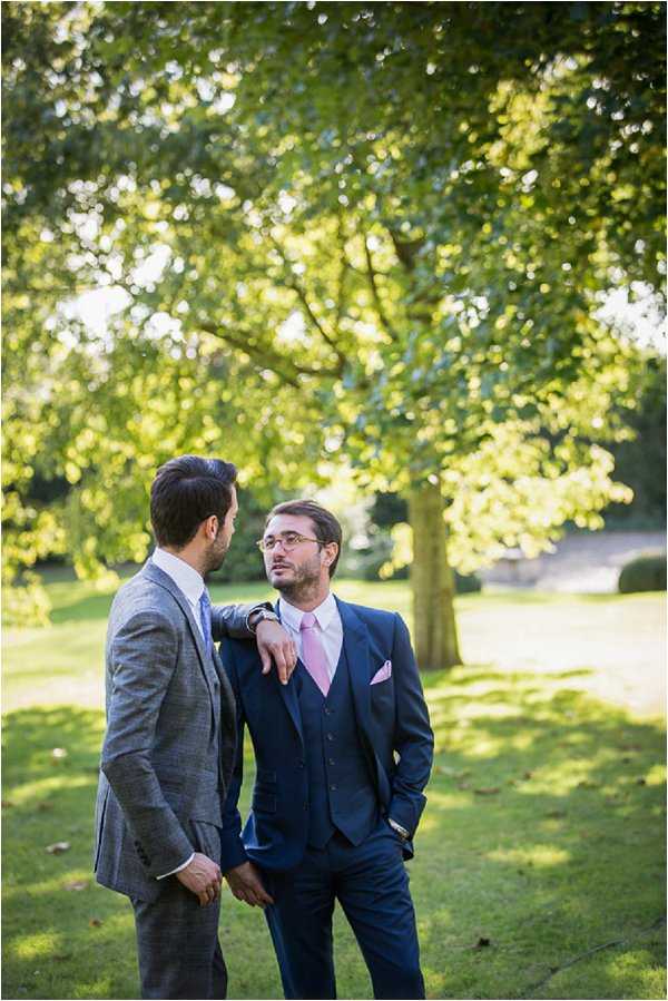 Two men stand together in a manicured garden setting, one resting his arm on the other's shoulder in a relaxed, candid pose. The man on the left wears a grey textured three-piece suit with a light blue tie, while the man on the right wears a navy three-piece suit with a pink tie and matching pink pocket square, paired with glasses. The shot is taken outdoors in a park-like garden with mature trees in soft focus behind them. The portrait is a mid-length shot with warm, natural late-afternoon light.