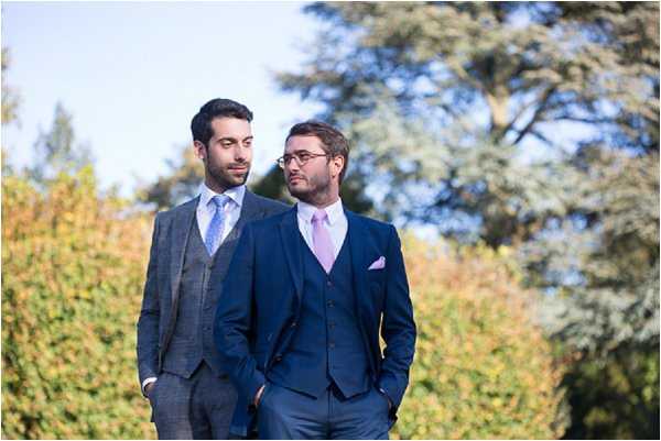 Portrait of two grooms standing outdoors in a garden setting, both with hands in pockets and gazing in the same direction. The groom on the left wears a charcoal grey three-piece suit with a light blue patterned tie, while the groom on the right wears a navy blue two-piece suit with a lavender tie and a pink pocket square. The styling is classic and coordinated, with complementary cool-toned accessories. The shot is a medium close-up portrait with a softly blurred green hedge background.