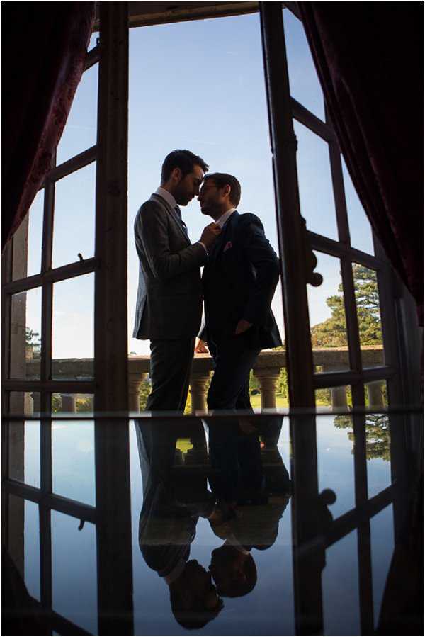 A couple portrait of two grooms standing close together in a doorway or large French window opening, their foreheads nearly touching as one adjusts the other's tie or lapel. Both men wear dark suits — one in charcoal grey with a pink pocket square, the other in navy — and are shown largely in silhouette against a bright blue sky. The highly polished reflective floor creates a near-perfect mirror image of the couple below them, adding strong visual symmetry to the composition. The setting appears to be a chateau or grand estate with a stone balustrade terrace and a ornamental pool or lake visible in the background through the open doors, with deep red curtains framing the interior sides of the frame. The shot is a full-length portrait taken from inside the venue looking outward, using backlighting to create a dramatic silhouette effect.