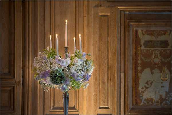 Close-up detail shot of a tall silver candelabra centerpiece holding five lit taper candles in blush pink, surrounded by a floral arrangement featuring soft lavender delphinium, blush roses, green hydrangea, baby's breath, and small trailing greenery. The piece is styled in a romantic, classic palette of blush, lavender, and soft green. The background shows warm honey-toned wood paneling and a partially visible ornate painted decorative panel with classical motifs, suggesting an interior room of a historic French chateau or manor house.