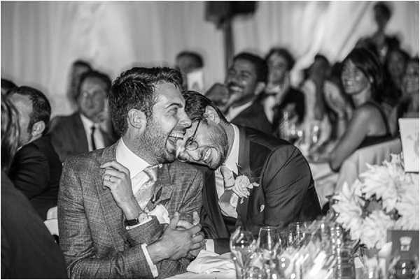 Black-and-white image captured during wedding reception speeches, showing two grooms seated at the head table laughing and leaning into each other with visible joy. One groom wears a checked suit jacket with a boutonniere, while the other wears a dark suit also with a boutonniere. In the foreground, glassware and what appear to be large-headed flowers such as dahlias are partially visible on the table. A crowd of approximately 15 or more guests is visible in the background, many also laughing and smiling. The image has strong contrast with bright highlights on faces and a soft, draped fabric backdrop. Medium close-up portrait composition.