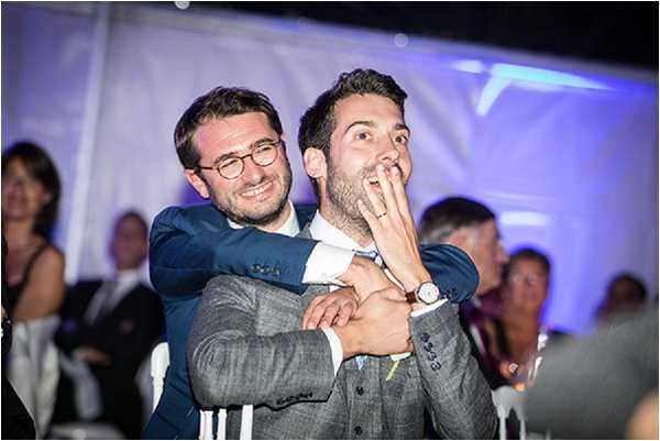 Two men share a candid, playful moment during what appears to be an indoor wedding reception held in a tent or marquee, lit with blue and purple ambient lighting. One man, wearing a navy suit with a bow tie and round glasses, is hugging the other from behind with a wide grin, while the second man, dressed in a grey checked suit with a light blue tie, reacts with a look of exaggerated surprise, his hand raised to his mouth. Several guests are visible and slightly out of focus in the background. The shot is a medium close-up with a shallow depth of field, capturing a spontaneous, lively reaction moment likely during speeches or an entertainment segment of the reception.
