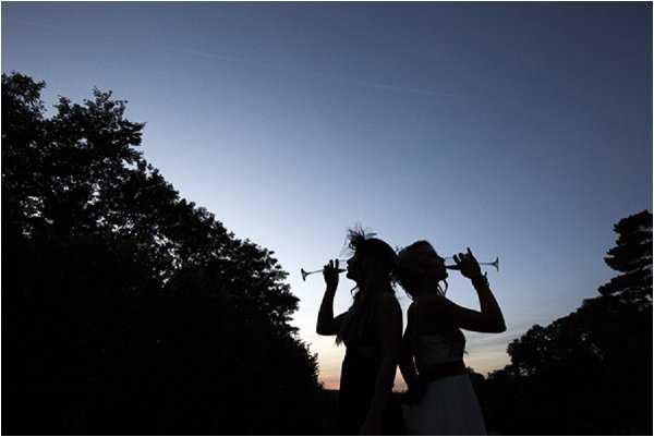 Two women, likely the bride and a guest or bridesmaid, are silhouetted against a dusk sky as they drink from champagne flutes, both tilting their glasses upward. The shot is taken outdoors at twilight, with deep blue tones in the sky and a faint warm glow at the horizon. The silhouettes show one figure wearing a hair accessory with feathering detail and the other with hair up. The composition is a wide portrait-style silhouette shot with trees framing both sides of the frame.