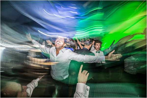 A heavily motion-blurred, wide-angle action shot of the wedding reception dance floor, taken with a slow shutter speed or zoom burst technique to create sweeping light trails in blue and green from the DJ or stage lighting. A man in a white dress shirt with his arms outstretched is laughing at the center of the frame, surrounded by other guests including one wearing a green bow tie and dark suit jacket. Multiple people are visible dancing energetically in the background. The intentional camera movement creates a chaotic, high-energy composition that emphasizes the lively atmosphere of the late-night reception party indoors.