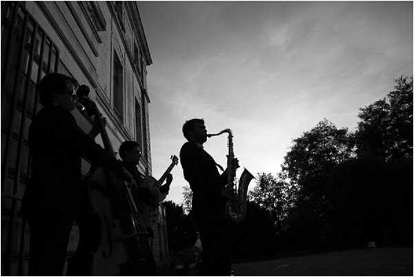 A black-and-white image of a live music ensemble performing outdoors at what appears to be a wedding cocktail hour or reception, positioned alongside the exterior wall of a large classical building. Three musicians are visible in strong silhouette against a bright sky, with identifiable instruments including a saxophone, a cello or double bass, and a guitar or smaller stringed instrument. The dramatic backlit composition renders the figures almost entirely in deep shadow with high contrast against the light background. Wide shot with a cinematic, atmospheric quality.