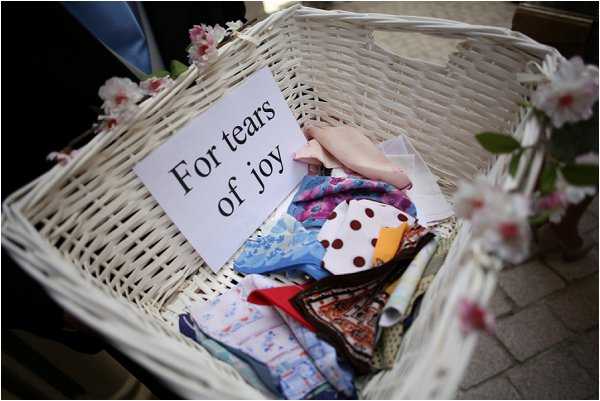 Close-up detail shot of a white wicker basket filled with assorted fabric handkerchiefs in various patterns and colors, including floral, polka dot, and patchwork designs in blues, reds, and pinks. A white card printed with the words 'For tears of joy' is propped inside the basket. The basket is decorated with small pink cherry blossom branches. This is a ceremony or pre-ceremony guest amenity detail, styled as a thoughtful touch for emotional guests.
