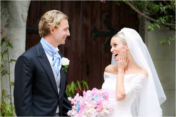 A couple portrait taken outdoors in front of a large dark wooden chapel door, likely just after the ceremony. The groom wears a dark navy suit with a light blue cravat and a white rose boutonniere, while the bride wears an off-the-shoulder white gown with long sleeves and a cathedral-length veil. The bride holds a loose, rounded bouquet of pink sweet peas, light blue hydrangeas, and pale pink blooms, and she is laughing with her hand raised to her cheek in a candid, joyful reaction. The shot is a medium two-person portrait with natural daylight illumination.