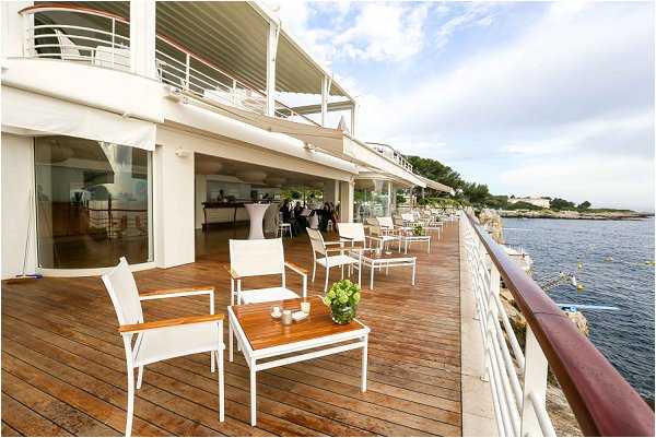 A wide shot of an outdoor waterfront terrace set up for a cocktail hour or pre-reception gathering. The venue features a multi-level modern white building with large glass doors opening onto a wooden deck that runs along the edge of a rocky coastline. White metal chairs and teak-topped side tables are arranged on the deck, with small green floral or plant centerpieces on the tables. In the background, guests and staff can be seen near tall white cocktail tables inside and just outside the venue space. The decor palette is minimal and clean, with white furniture and natural wood tones. Potential venue feature image.