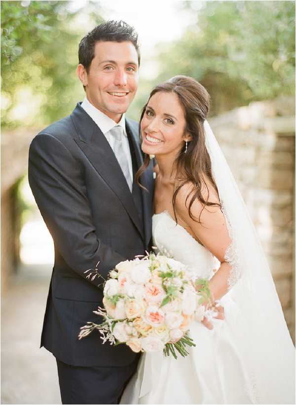 A couple portrait taken outdoors, likely in a courtyard or garden setting with soft natural backlighting. The bride wears a strapless white gown with beaded detailing along the back neckline and a cathedral-length veil, with her dark hair styled in a half-up, half-down look; she holds a rounded bouquet of blush and peach garden roses with green foliage. The groom wears a dark charcoal suit with a light grey tie and white dress shirt. The composition is a close-up portrait with a shallow depth of field, blurring the background foliage and stone archway elements behind the couple.