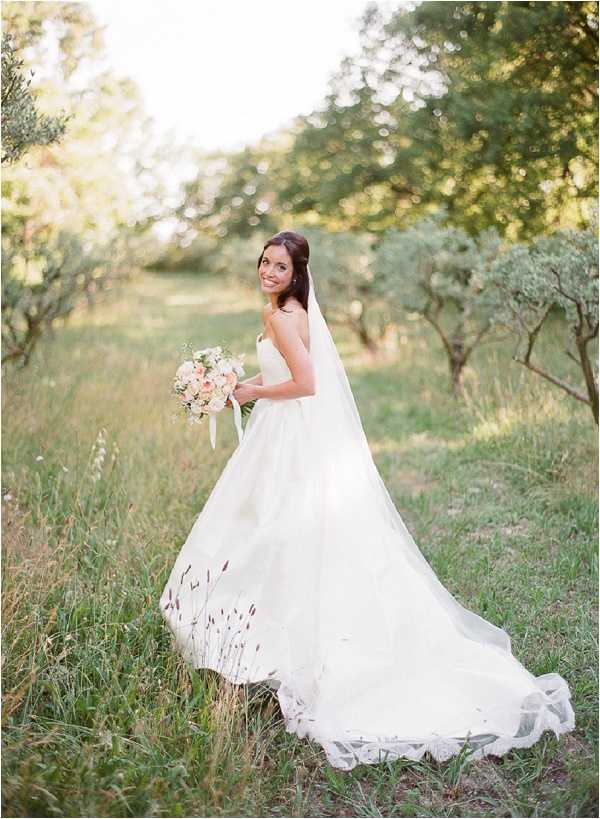 A bridal portrait taken outdoors in what appears to be an olive grove or orchard setting, with the bride standing alone and turning back toward the camera with a smile. She wears a strapless ivory ballgown with a full skirt and an extremely long cathedral-length veil that trails across the grass behind her. The gown features delicate floral embroidery or appliqué detail along the hem and train. She holds a loosely gathered bouquet of blush pink roses, white blooms, and greenery tied with an ivory ribbon. The shot is a full-length portrait with soft, warm backlighting and a shallow depth of field that blurs the trees in the background.