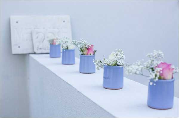 Detail close-up of a wedding decor arrangement featuring five small periwinkle blue ceramic pots lined up on a white surface, each containing mini bouquets of pink and white roses with white gypsophila. In the background, slightly out of focus, is a white decorative sign or plaque with raised lettering, likely displaying the wedding date or couple's initials. The overall color palette is soft and muted — periwinkle blue, blush pink, and white — suggesting a pastel-themed wedding. The setting appears to be indoors against a light grey wall.