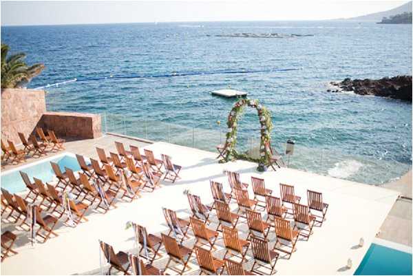 An outdoor wedding ceremony setup photographed from an elevated wide-angle perspective, showing rows of wooden folding chairs arranged in two sections on a white terrace directly overlooking the sea. A circular floral arch decorated with greenery and colorful blooms stands at the altar position, positioned against a glass balustrade with open water as the backdrop. The setting appears to be a modern coastal venue terrace adjacent to a pool, with no guests or couple present — the space is arranged and ready for the ceremony. The overall decor style is clean and modern with natural wooden chairs and a lush floral arch as the focal point. Potential venue feature image.