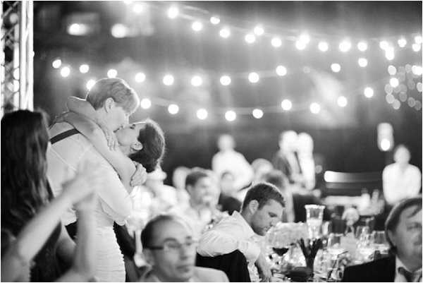 Black-and-white image of a wedding reception scene, shot indoors under strings of globe fairy lights strung across the ceiling. The couple stands and shares a kiss in the left-center of the frame, the bride in a fitted strapless dress and the groom in a white shirt with suspenders, embracing closely. Surrounding them, approximately 15–20 seated guests are visible at round dinner tables set with glassware, some watching the couple and others engaged in conversation. The fairy lights create bright bokeh circles in the background, adding strong contrast against the darker reception space. Medium-wide portrait-style shot with shallow depth of field that keeps the kissing couple in focus while the background guests are softly blurred.
