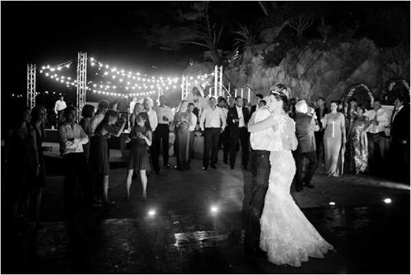 Black-and-white image of a couple's first dance taking place outdoors at night on a polished dance floor, surrounded by approximately 30-40 guests watching from the perimeter. The bride wears a fitted lace mermaid-style gown and the groom is in a dark suit. String bistro lights are strung between metal truss structures overhead, providing the primary ambient lighting along with small floor-level uplights marking the dance floor edge. The rocky natural backdrop and palm trees suggest a clifftop or coastal venue setting. The wide shot captures the full scene with guests in formal attire, some holding drinks, arranged in a semicircle around the dancing couple.