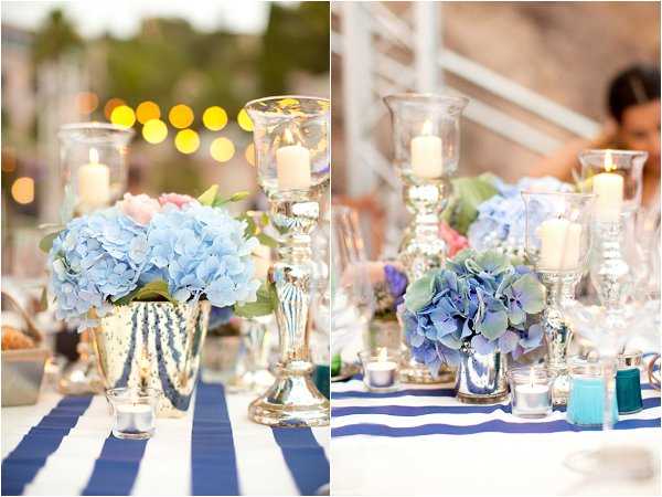 Close-up detail shots of wedding reception table centerpieces styled in a nautical theme. The tables are dressed with navy blue and white wide-stripe linens, and the centerpieces feature light blue and periwinkle hydrangeas arranged in mercury glass vases. Tall silver mercury glass candlesticks hold glass hurricane candle holders with white pillar candles, and small glass votive holders are scattered across the table. Soft bokeh lights are visible in the background of the left image, and a guest is partially visible in the background of the right image. The overall decor palette is navy, silver, and powder blue with accents of blush pink florals.