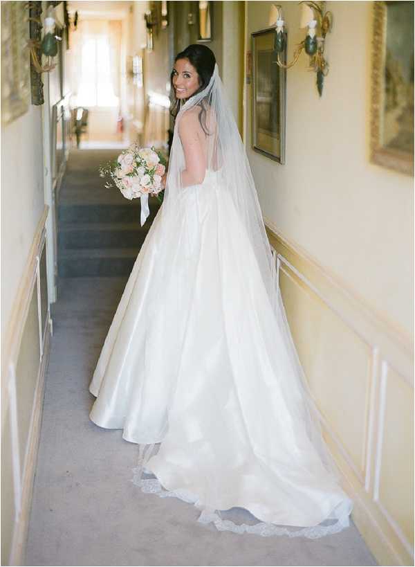 A bridal portrait taken indoors in a hallway of what appears to be a chateau or manor house, decorated with wall sconces, framed paintings, and paneled wainscoting in pale yellow tones. The bride stands with her back to the camera and glances over her shoulder, wearing a full-skirted ivory satin ball gown with an open back and a cathedral-length veil trimmed with lace edging. She holds a rounded bouquet of blush and soft pink roses with small white filler flowers. The composition is a three-quarter portrait emphasizing the back of the gown and the trailing veil, with natural light coming from a window at the far end of the corridor.