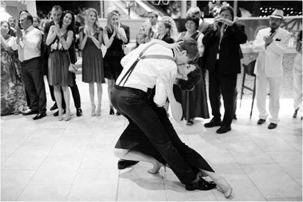 A black-and-white image of a couple sharing a deep dip and kiss on the dance floor during what appears to be their first dance at a wedding reception. The groom is dressed in dark trousers, a white dress shirt, and suspenders, while the bride wears a full-length dark gown and is barefoot. Approximately 15–20 guests are visible in the background forming a circle around the dance floor, many clapping and reacting with enthusiasm. The reception appears to be held in a covered outdoor or semi-open venue with tiled flooring, and a musician with a brass instrument is visible in the background. The image is a medium-wide shot with strong contrast between the bright dance floor and the darker background.