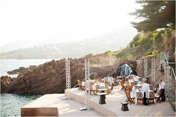 Wide shot of an outdoor wedding reception setup on a coastal terrace positioned directly at the water's edge, with dramatic red rock formations and the sea visible in the background. The reception space features round and rectangular tables dressed in white linens, paired with natural wood folding chairs, arranged across a smooth stone terrace. String lights are strung between metal truss poles across the space, and a few staff members in white shirts are visible attending to the tables. The overall decor palette is clean and minimal — white, natural wood, and black accents — giving the setup a modern, understated feel. Potential venue feature image.