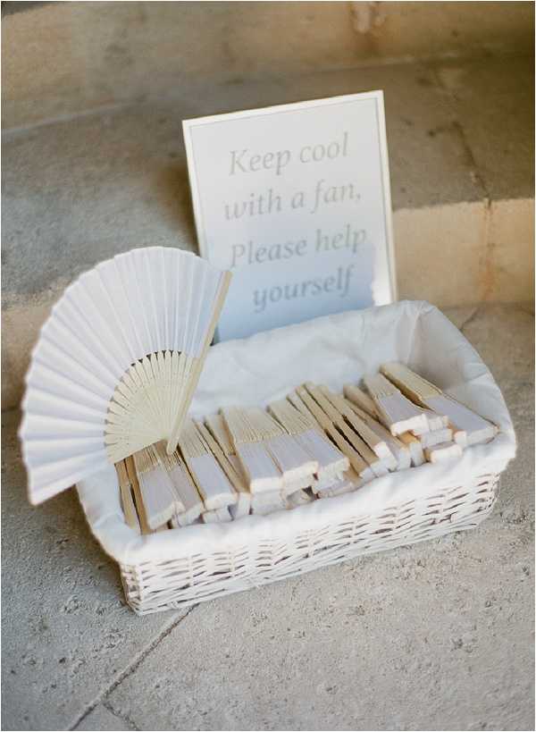 Close-up detail shot of a guest amenity station featuring a white wicker basket filled with folded white and natural wood hand fans, lined with white tissue paper. One fan is displayed open to show its full shape. A printed card in the basket reads 'Keep cool with a fan. Please help yourself' in soft grey lettering on a white background. The setup is placed on a stone floor, suggesting an outdoor or semi-covered ceremony or cocktail area. The overall styling is clean and minimal with an all-white and natural wood palette.