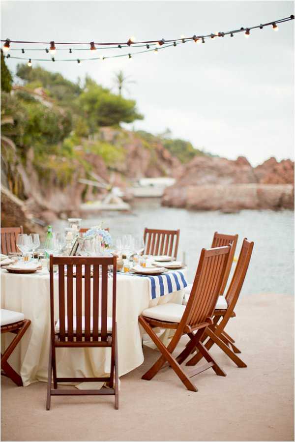 An outdoor wedding reception table set directly beside the water at a coastal location with red rocky cliffs and green vegetation in the background. The round table is dressed with a cream linen tablecloth and a bold blue-and-white striped table runner, set with white plates, wine glasses, and a small centerpiece of light blue flowers. Dark-stained wooden folding chairs with cream cushions surround the table. Overhead, a string of globe Edison-style bulb lights runs across the frame, adding warm ambient lighting to the nautical-themed tablescape. Wide shot capturing the full table setting and waterside backdrop.