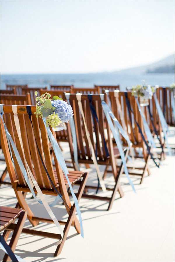 Detail shot of an outdoor wedding ceremony seating arrangement set on a terrace or deck overlooking the sea. Wooden folding chairs are arranged in rows and decorated with trailing light blue and navy ribbons, along with small glass vessels holding blue hydrangeas and green foliage tied to the aisle-side chairs. The decor palette is nautical, combining navy, pale blue, and natural wood tones. The composition is a shallow depth-of-field portrait-orientation shot focused on the nearest aisle chair, with the remaining rows receding into a soft blur against a coastal backdrop.
