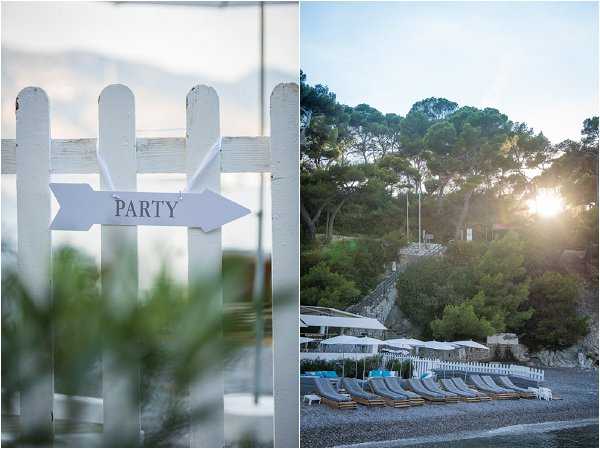 A diptych image showing two detail shots of an outdoor coastal wedding venue. The left panel is a close-up of a white picket fence with a white arrow-shaped sign reading 'PARTY' attached to it, with soft bokeh greenery in the foreground. The right panel is a wide shot of a beach club venue at golden hour, showing a row of grey sun loungers with white umbrellas arranged along a pebble beach, backed by a white picket fence, white parasols over a terrace area, and pine trees on a hillside with sunlight breaking through. The venue has a casual coastal aesthetic with a white and grey color palette. Potential venue feature image.