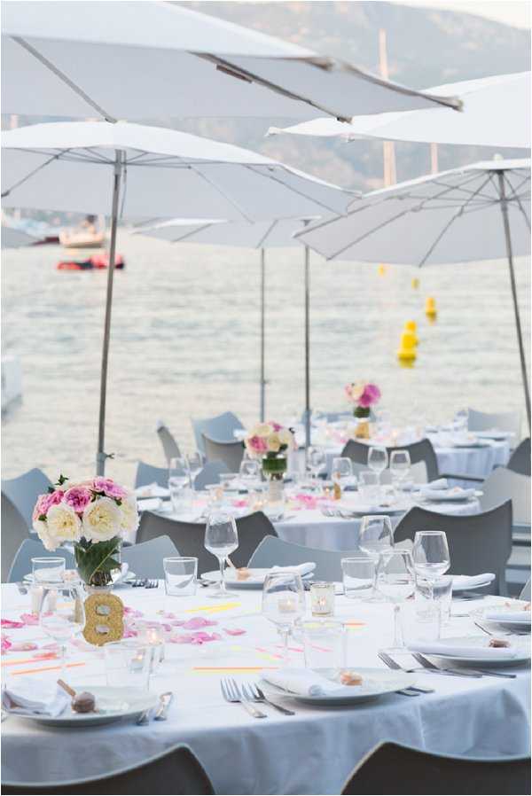 An outdoor waterfront wedding reception setup, photographed in a wide portrait composition showing multiple round dining tables arranged directly beside the sea. The tables are dressed in white floor-length linens and set with white plates, folded white napkins, silver cutlery, and an assortment of wine and water glasses. Centerpieces consist of clear vases holding loose arrangements of cream and pink garden roses and peonies, with scattered pink flower petals and small neon yellow and pink paper accents spread across the tabletops. A gold glitter table number marker is visible on the foreground table. Small votive candles are placed between the glasses. The chairs are modern gray molded chairs. Large white market umbrellas shade the tables, and the overall decor palette is white, pink, and neon yellow with a modern, clean aesthetic. No guests are present, suggesting this was taken during setup or before guests were seated.