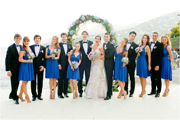 A bridal party group portrait taken outdoors at a coastal or lakeside venue, with a hillside visible in the background. The couple stands at the center — the bride in a cap-sleeve lace gown in ivory with subtle floral detailing, and the groom in a dark navy suit with a bow tie and pink boutonniere. The bridal party consists of approximately five bridesmaids in cobalt blue knee-length wrap-style dresses carrying small mixed bouquets of pink, blue, and lavender blooms, and approximately five groomsmen in black tuxedos with bow ties and white boutonnieres. A circular floral arch frames the couple in the background, decorated with blue hydrangeas, pink roses, and greenery. The shot is a wide group portrait taken at eye level in natural daylight.