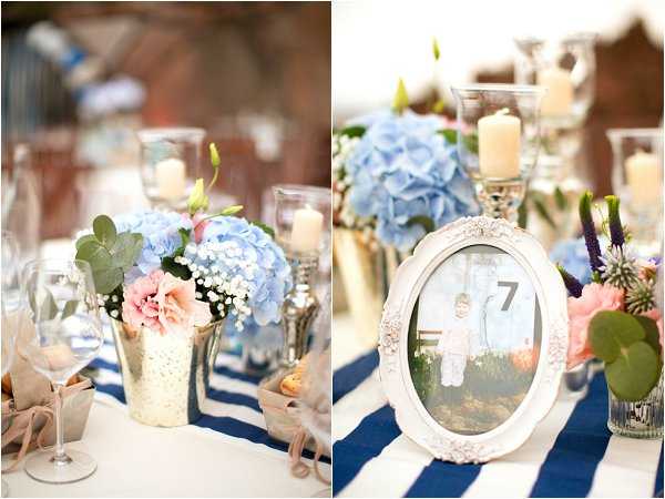 Close-up detail shots of wedding reception table decor, shown as a diptych. The left image features a gold mercury glass vase holding light blue hydrangeas, blush pink lisianthus, and white baby's breath alongside eucalyptus greenery, with pillar candles in glass hurricane holders and wine glasses visible on the table. The right image shows a white ornate oval picture frame used as a table number marker, displaying the number 7 alongside a small photo, with blue hydrangeas, blush pink florals, purple thistle, and candles in the background. The table is dressed with a navy and white wide-stripe table runner. The overall decor palette combines navy blue, blush pink, light blue, and gold accents in a classic-with-nautical-touches style. Both are detail/close-up shots with a shallow depth of field.