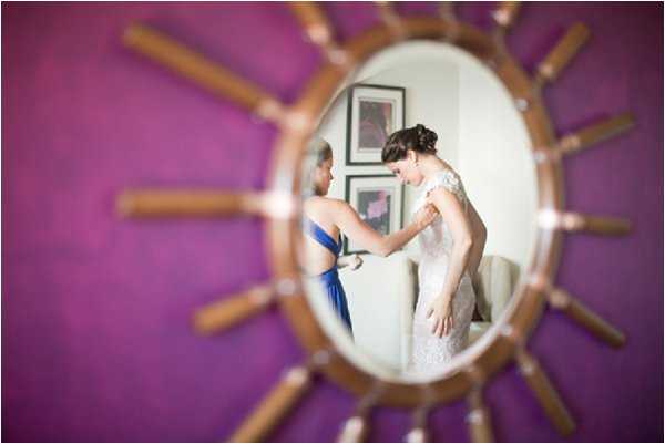 A getting-ready scene captured as a reflection in a sunburst-style oval mirror with a wooden spoke frame, mounted on a vibrant magenta-purple wall. The reflection shows a bride in a fitted lace gown with cap sleeves having her dress fastened by a helper dressed in a cobalt blue strapless dress. Two framed artworks are visible on the white wall behind the pair in the reflection. The composition uses the mirror as a creative framing device, with the mirror itself in sharp focus while the surrounding room is intentionally blurred, creating a portrait-style detail shot.