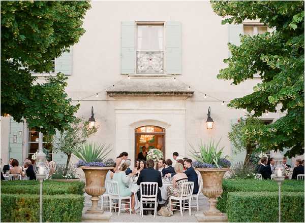 An outdoor wedding reception is taking place in a formal garden courtyard directly in front of a classic French manor house with pale cream render and sage green shutters. Approximately eight to ten guests are seated around a round table dressed in white linens, with a large white floral centerpiece visible at its center, while additional guests are visible at tables to the sides. The garden features neatly clipped low boxwood hedges, large stone urns planted with agave and purple flowering plants, and string lights are visible above the entrance doorway. The setting is classic French countryside in style, photographed as a wide shot during early evening with warm ambient light from wall-mounted lanterns flanking the manor entrance. Potential venue feature image.