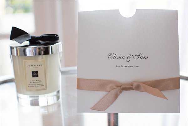 A close-up detail shot of two wedding styling items placed on a reflective glass surface. On the left is a Jo Malone London candle in a clear glass vessel with a silver lid and a black satin bow. On the right is a white wedding stationery booklet printed with 'Olivia & Sam' and the date '6th September 2014' in black script font, tied with a blush pink satin ribbon bow. The composition is a horizontal detail/flat-lay style image with a soft, bright background.
