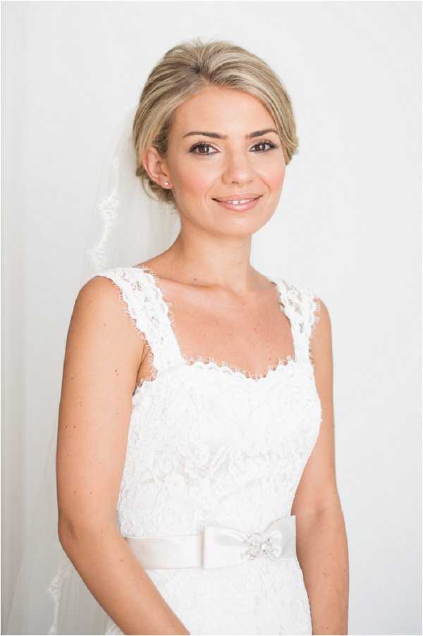 A close-up portrait of a bride photographed against a plain white background, likely taken during the getting-ready phase. She wears a white lace wedding dress with wide lace straps, a square neckline, and a white satin ribbon sash at the waist featuring a small crystal brooch detail. Her shoulder-length blonde hair is styled in a low updo, and she wears a plain-edged white veil falling behind her shoulders. Her makeup is natural and polished, featuring neutral tones, defined brows, and pearl stud earrings.