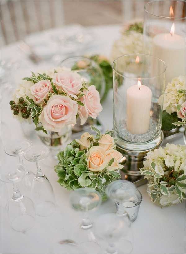Close-up detail shot of a wedding reception table centerpiece arrangement on a white linen tablecloth. The centerpiece features multiple small low-profile floral clusters with blush pink roses, peach roses, cream hydrangeas, green hydrangeas, variegated ivy, and green berry stems arranged in glass vases on silver pedestals. Two lit pillar candles in tall clear glass hurricane holders with small glass pebbles at the base are interspersed among the florals. Several empty wine glasses are tipped on their sides near the arrangement, and small glass tealight holders are scattered across the table surface. The overall decor palette is blush, peach, cream, and green with silver accents, giving the tablescape a classic, garden-style aesthetic.