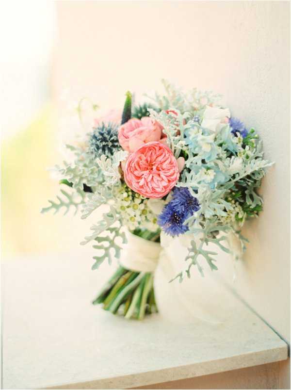 Close-up detail shot of a bridal bouquet resting on a light stone or marble ledge against a pale wall. The bouquet features coral-pink garden roses as the focal blooms, paired with pale blue hydrangeas, violet-blue cornflowers, dusty miller foliage with its silvery-grey lobed leaves, small white wax flower clusters, and touches of deep purple anemone. The stems are wrapped in wide ivory satin ribbon tied in a loose bow. The overall palette is soft and muted — coral, periwinkle, pale blue, and silvery-green — suggesting a romantic, garden-style floral design. The image is shot with a shallow depth of field and slightly soft focus, consistent with a film photography aesthetic.