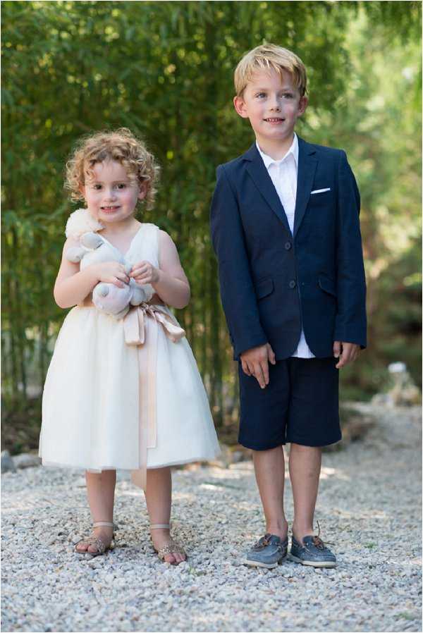 Portrait of two young children dressed for a wedding, standing side by side on a gravel path with lush green bamboo in the background. The flower girl wears a knee-length ivory tulle dress with a blush pink satin sash tied at the waist, silver sandals, and holds a small stuffed animal toy; the page boy wears a navy blazer with a white pocket square, a white shirt, navy shorts, and grey boat shoes. Both children face the camera directly in a classic upright portrait composition shot outdoors in natural daylight.