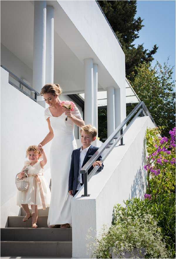 A bride descends an outdoor staircase of a modern white villa or venue, accompanied by two young children acting as flower girl and page boy. The bride wears a slim-cut white dress with short flutter sleeves and carries a small bouquet of pink and coral flowers. The flower girl wears a cream dress with a blush sash and carries a white wicker basket, while the page boy wears a navy suit with a pink tie. The setting is a contemporary white rendered building with columned upper terrace and metal railings, shot as a medium full-length portrait from a slightly lower angle.