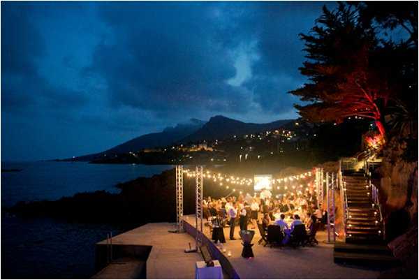 An outdoor evening wedding reception taking place on a coastal terrace or platform directly beside the sea, with a mountainous coastline and town lights visible in the background. Approximately 50-80 guests are seated at round tables dressed in purple or blue linens, lit by a canopy of warm bistro/festoon string lights suspended from metal truss structures. The reception area is accessed via stone or concrete steps on the right side of the frame, with a large tree lit by red uplighting adding a dramatic accent. The wide-angle aerial shot captures the full scope of the venue setup against the deep blue dusk sky and waterfront setting. Potential venue feature image.