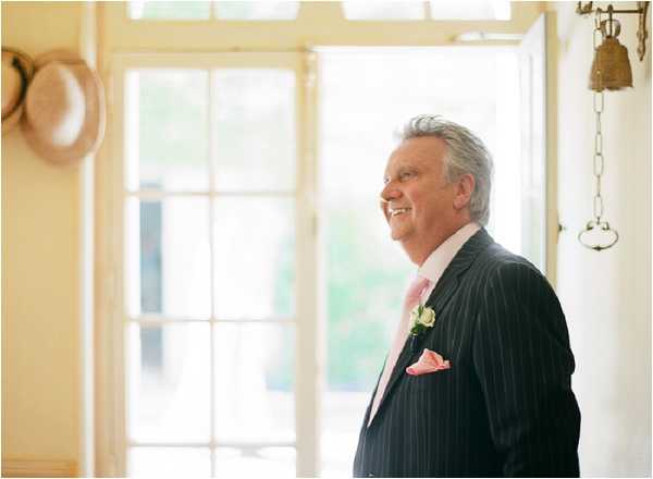 A man, likely the father of the bride or groom, stands smiling inside a bright room with French doors open behind him, suggesting a chateau or country house venue. He is dressed in a dark charcoal pinstripe suit with a pale pink tie, a pink pocket square, and a white boutonnière with a small pink accent flower. A straw hat hangs on the wall to the left, and a decorative brass bell on a chain is visible to the right. The image is a medium portrait shot with soft, natural backlit lighting coming through the open doors.