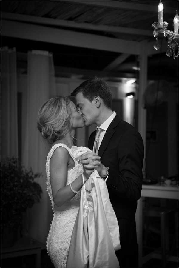 This black-and-white portrait captures a bride and groom sharing a kiss at what appears to be an evening reception. The bride wears a fitted lace dress with cap sleeves and holds what appears to be a jacket or wrap in her hands, while the groom is dressed in a dark suit with a light-toned tie. The setting is a covered outdoor or semi-enclosed terrace with a wooden pergola ceiling, white curtain draping visible in the background, and a crystal chandelier with candle-style lights in the upper right corner. The image has strong contrast with deep shadows and warm ambient lighting glowing from the interior space behind the couple, shot as a close-up portrait with a shallow depth of field.