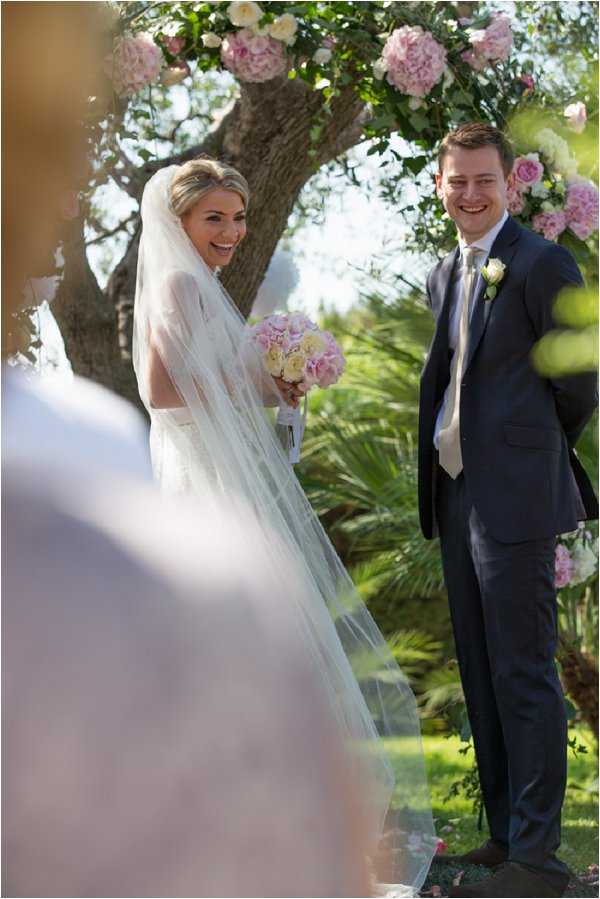 An outdoor wedding ceremony captures the bride and groom standing at the altar beneath a floral arch decorated with blush pink and cream peonies and roses. The bride wears a white fitted gown with a long cathedral veil and holds a bouquet of soft pink and ivory roses and hydrangeas, while laughing and turning slightly toward the camera. The groom wears a navy suit with a cream tie and a small white rose boutonniere, smiling broadly. The setting is a garden with palm and olive trees visible in the background, and a blurred guest figure appears in the foreground, shot from a slightly angled perspective.